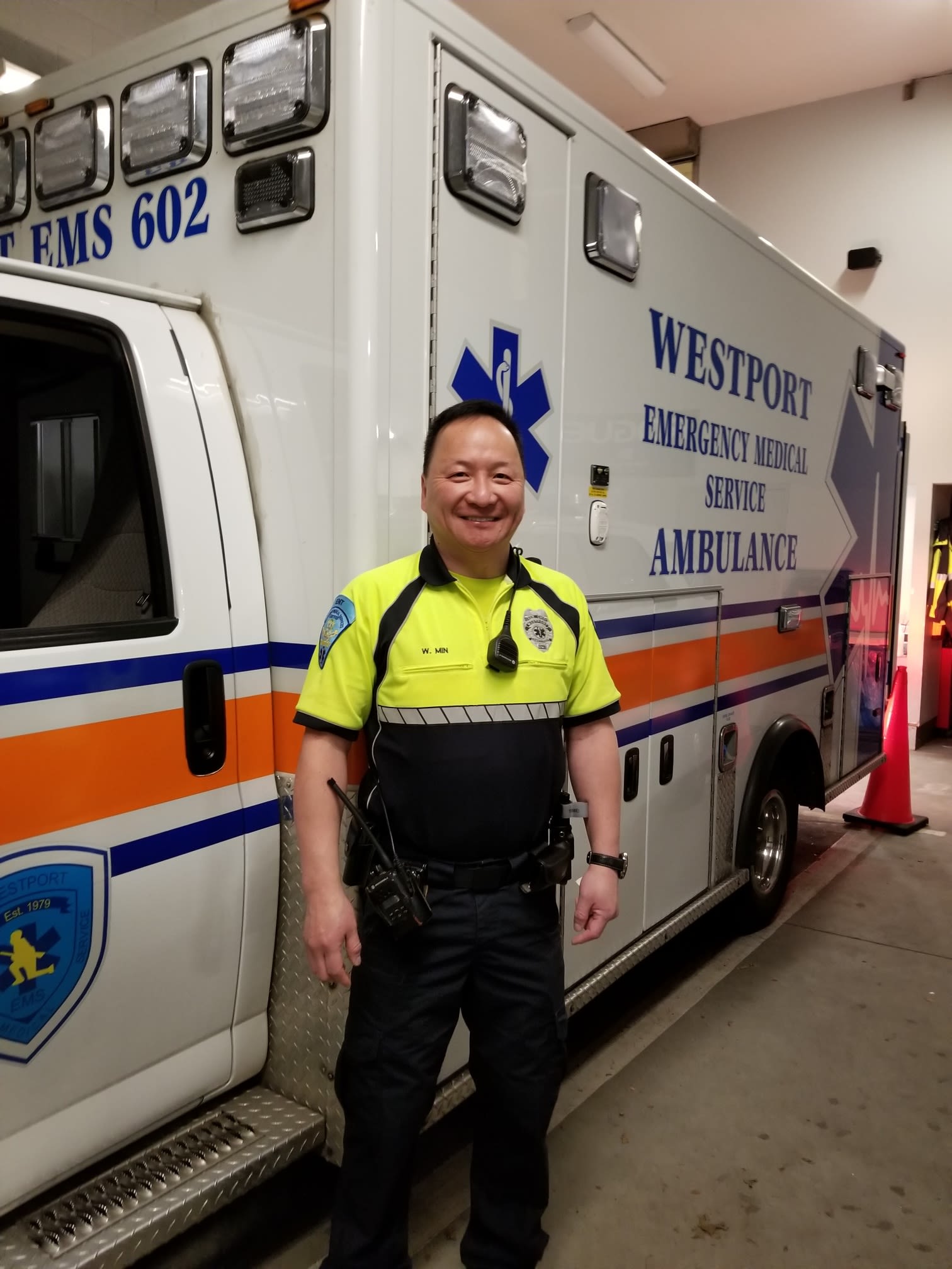 Mr. Bill Min in his EMT uniform standing next to the Westport Emergency Medical Services Ambulance.